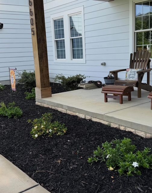 Front porch with white siding, wooden post, chairs, and dark mulch landscaping by a concrete walkway