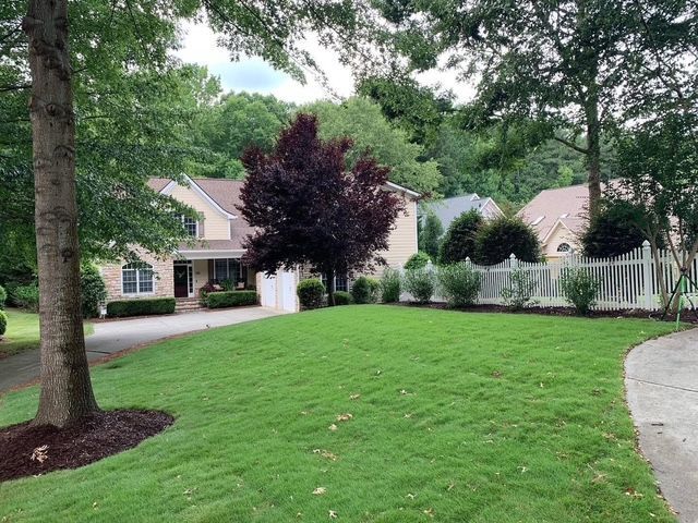 Suburban front yard with green lawn, trees, curved driveway, and white houses in the background