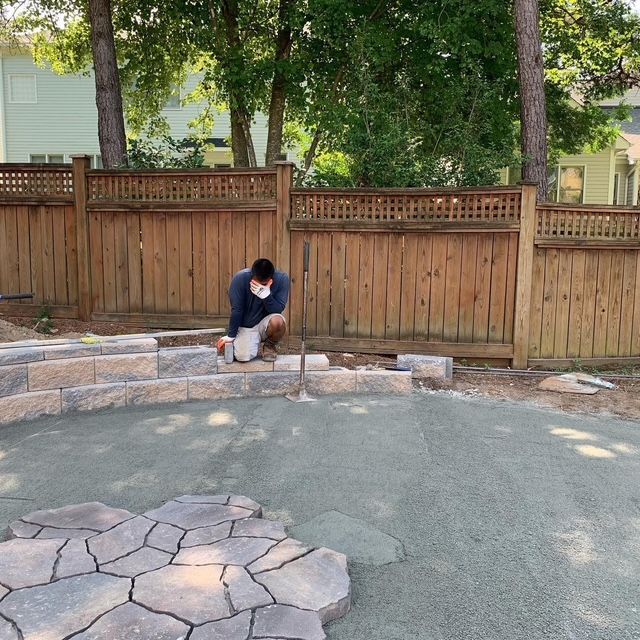 Person kneeling by a curved stone edging in a backyard with a wooden fence and trees.
