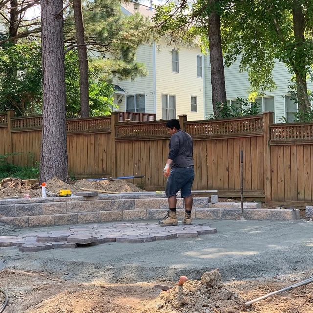 Worker standing in a backyard near a stone fire pit, wooden fence, and trees.