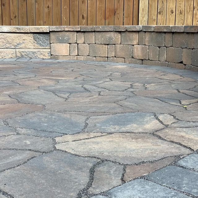Stone patio with irregular flagstones and a curved stacked-stone wall beneath a wooden fence.
