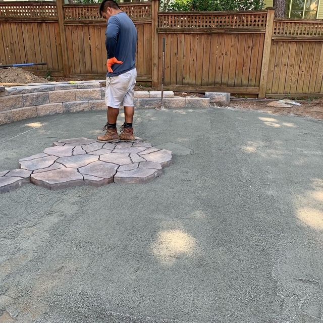 Person standing on a stone patio in a fenced backyard, with a raised stone border and wooden fence behind.