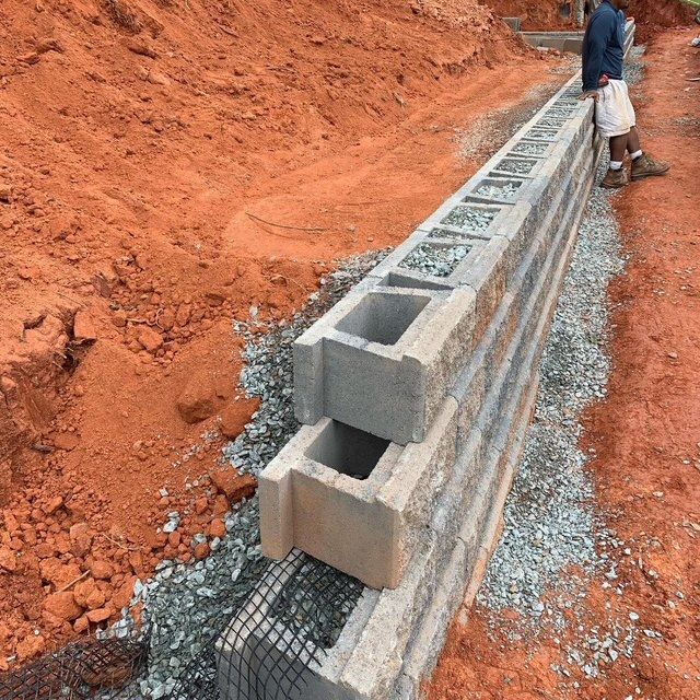 Concrete block retaining wall under construction beside a red dirt slope, with workers at the top.