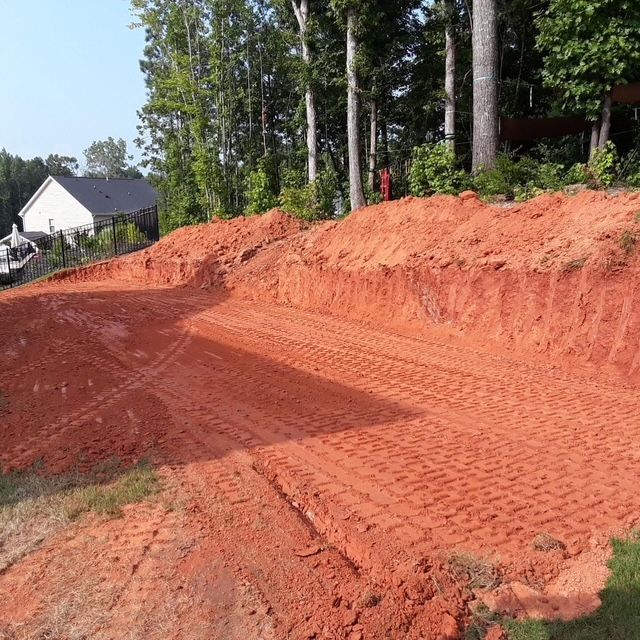 Freshly graded red dirt road beside a wooded area, with a white house in the background.