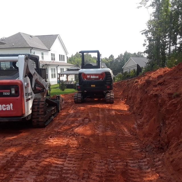 Two Bobcat excavators on a muddy construction road beside a white house and red dirt embankment.