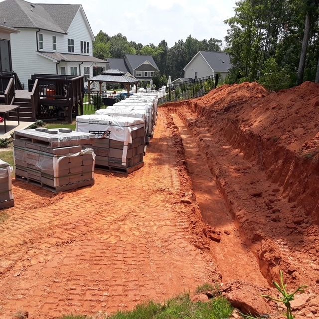 Construction materials beside a newly dug red dirt trench along a suburban street near houses.