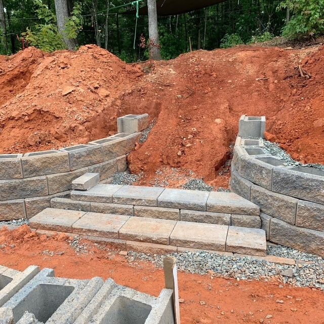 Stone retaining wall and steps under construction, with red dirt embankment behind them.