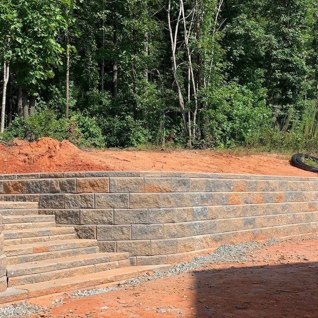 Tiered stone retaining wall with steps on a red dirt slope beside a wooded area