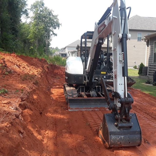 Excavator grading a red dirt slope beside a suburban house and driveway