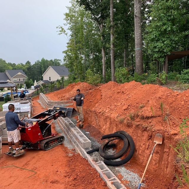 Workers loading a red mini skid steer in a dirt trench beside a retaining wall under construction