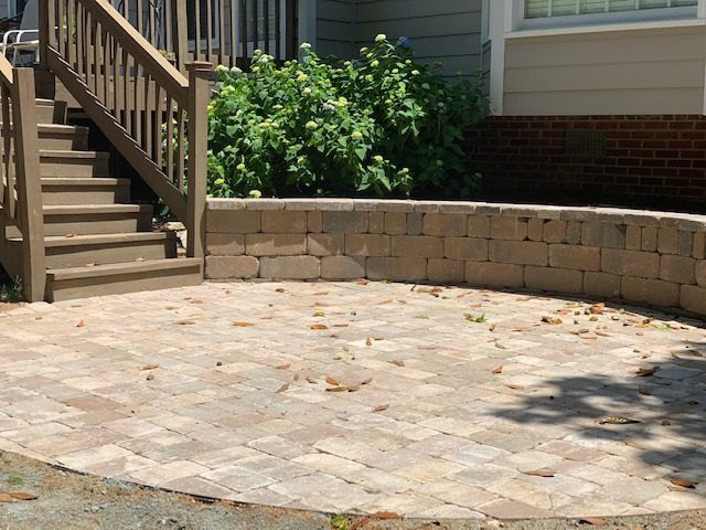 Stone patio with curved retaining wall, stairs, and green shrubs beside a house
