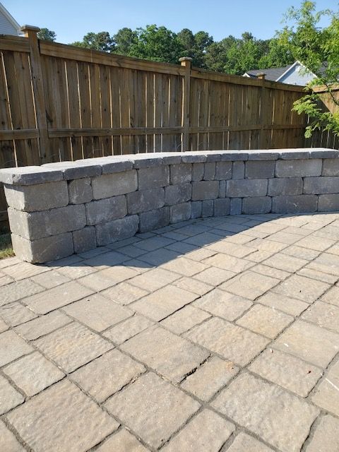 Curved gray stone retaining wall along a paved patio beside a wooden fence in a sunny backyard
