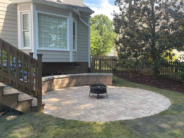 Backyard patio with a circular stone площадка, fire pit, steps, and a house exterior with fence and trees.