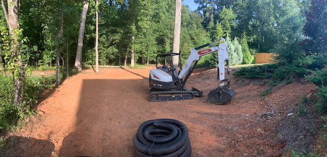 Excavator on a red dirt road in a wooded area with stacked tires in the foreground