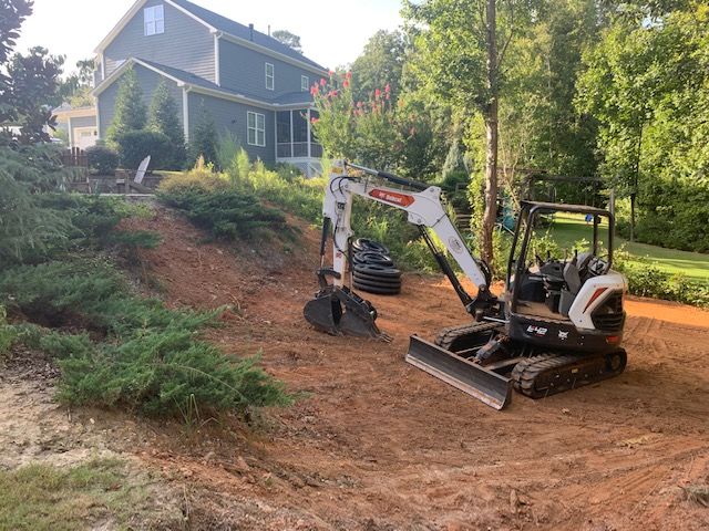 Compact excavator leveling a red dirt slope beside a house and trees