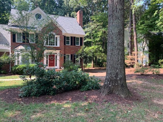Red-brick two-story house with white trim and large trees in a wooded yard
