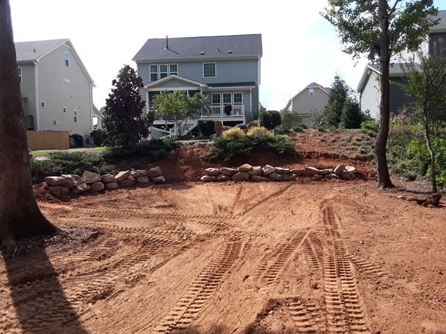 Dirt driveway under construction leading to a suburban house with rock retaining wall and trees