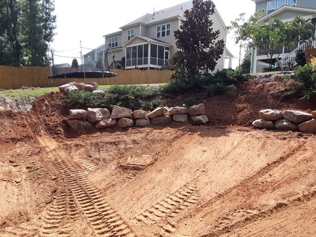 Backyard excavation with a dirt slope, rock retaining wall, and large house in the background