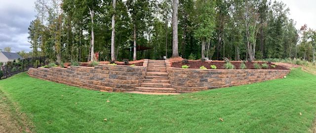 Wide grassy lawn before a terraced stone retaining wall with trees and shrubs in the background.