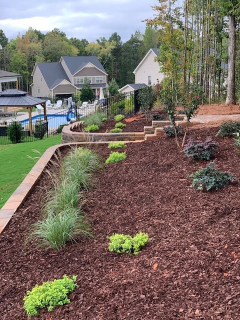 Mulched landscaped slope with stone steps, shrubs, and a poolside suburban neighborhood in the background