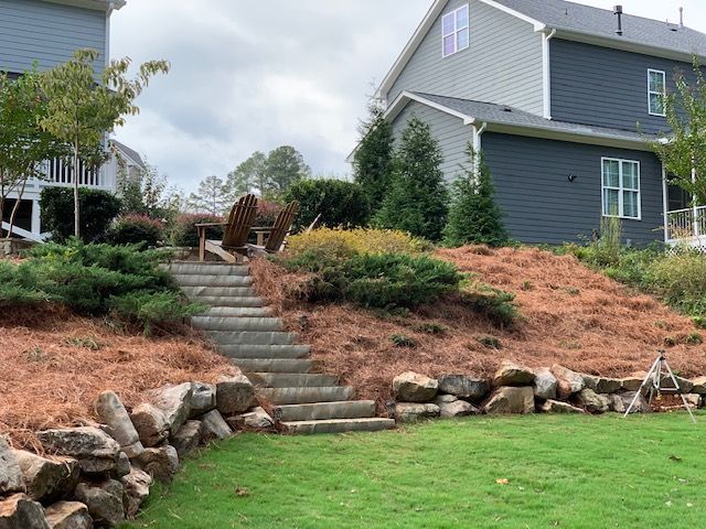 Landscaped backyard with stone steps, retaining wall, green lawn, and gray house on a hillside.