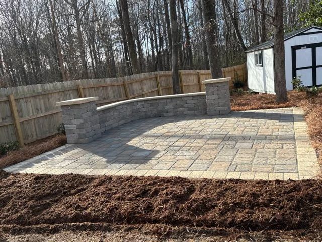 Patio with stone pavers and a curved retaining wall in a backyard, bordered by fresh mulch and a fence