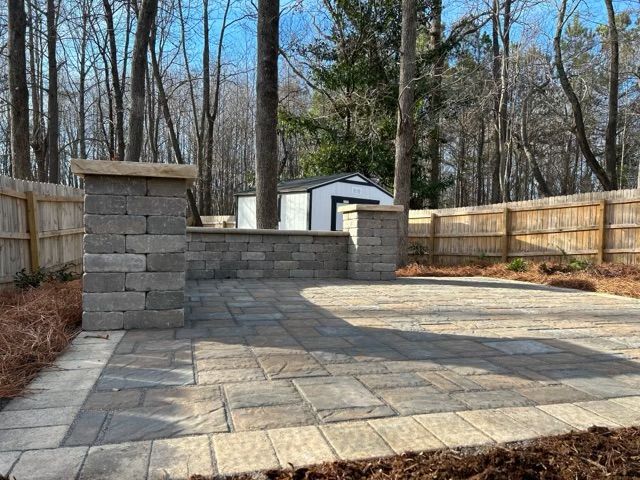 Stone patio with low retaining wall in a fenced backyard, trees and a shed in the background