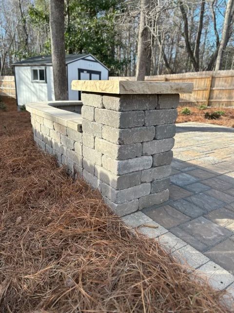 Stone retaining wall beside a paved patio, with a small shed and leafless trees in the background