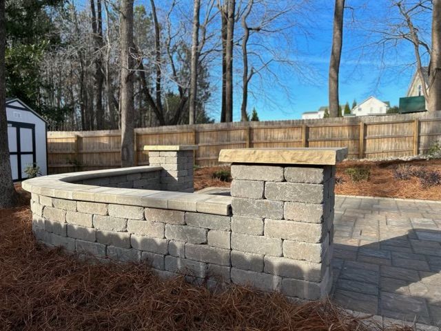 Stone garden wall and curved retaining wall beside a paved path in a backyard, with trees and fence in the background