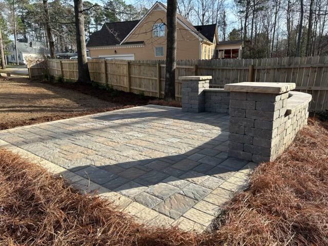 Paver patio with stone steps and retaining walls in a fenced backyard, with a tan house in the background