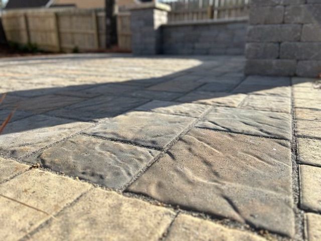 Stone patio with a curved walkway in sunlight, bordered by a low stone wall and a wooden fence.