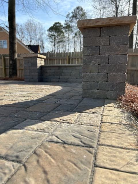 Stone patio with a brick pillar and low wall beside a wooden fence in a sunny yard