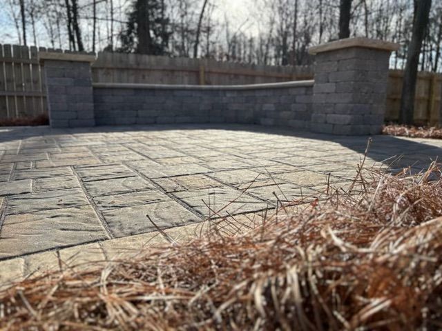 Stone patio with low retaining wall and bare trees, viewed from ground level behind dry grass.