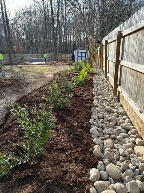Mulched garden bed with young plants beside a wooden fence and river rocks, in a yard under construction
