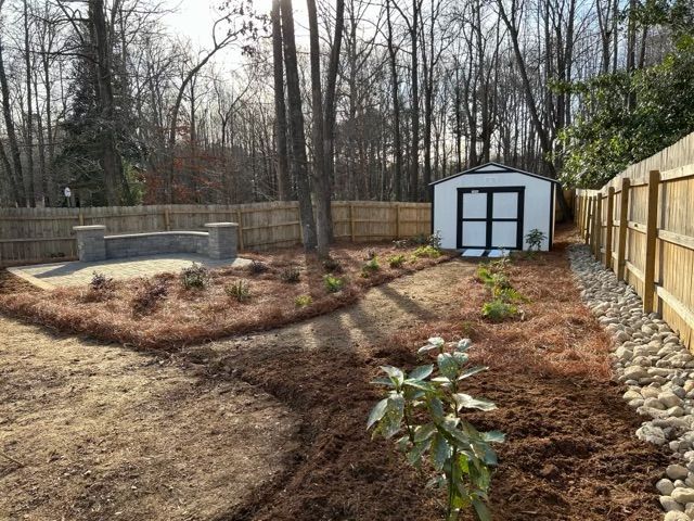 Backyard garden with mulched beds, trees, a stone patio, and a small white shed by a wooden fence