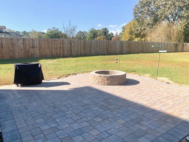 Backyard patio with pavers, fire pit, black grill, and fenced lawn under blue sky
