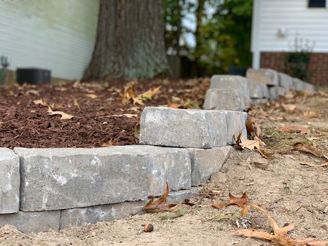 Tiered gray stone border around a tree bed with mulch and scattered leaves beside a house