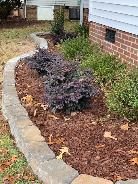 Curved stone garden border with purple shrubs and mulch beside a brick house.