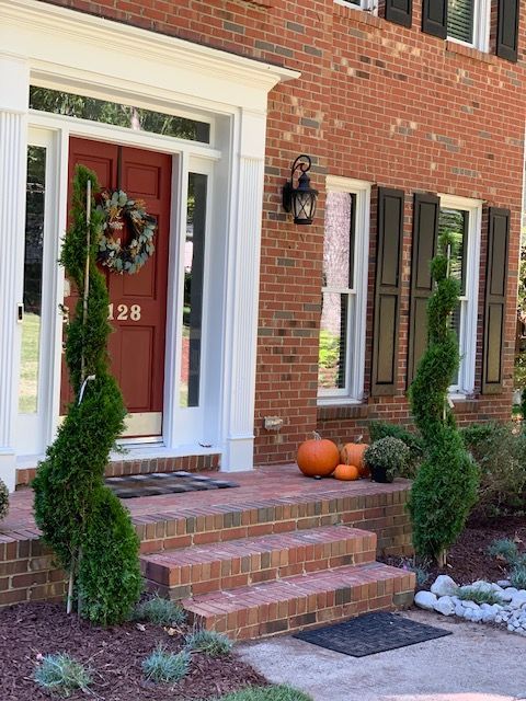 Brick house front porch with red door, fall wreath, topiary planters, and pumpkins on the steps