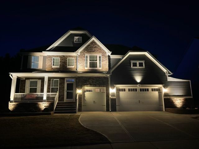 Nighttime view of a house front lit by porch lights, with a small tree and a detached garage.
