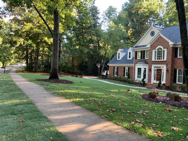 Brick suburban house with white trim and a tree-lined front yard beside a concrete walkway
