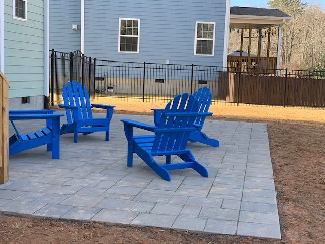 Blue Adirondack chairs on a stone patio beside a house and fenced yard.