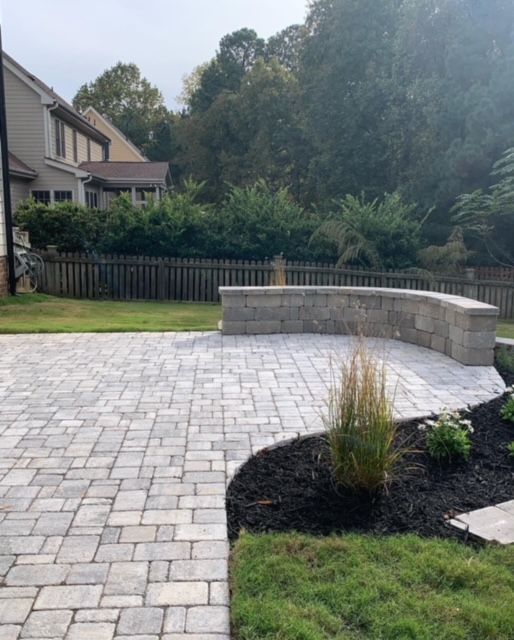 Backyard patio with gray pavers, curved stone bench, grass, and trees beside a house