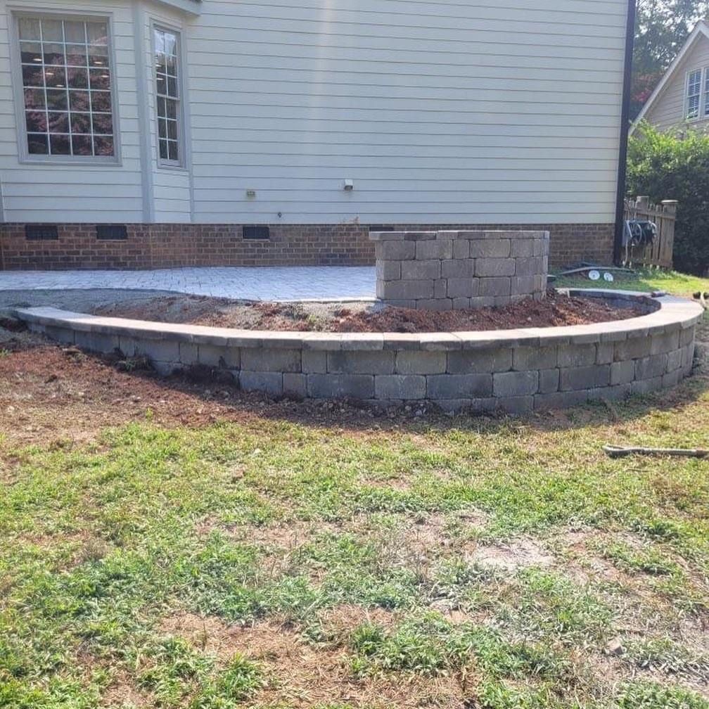 Curved stone retaining wall and raised patio beside a house, with patchy grass in the foreground.