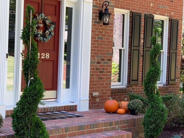 Front porch with pumpkins, wreath on red door, and two tall spiral topiary plants beside brick columns