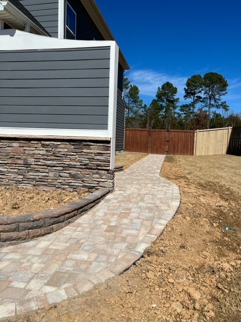Curved paver walkway beside a gray house leading to a wooden fenced backyard under a blue sky