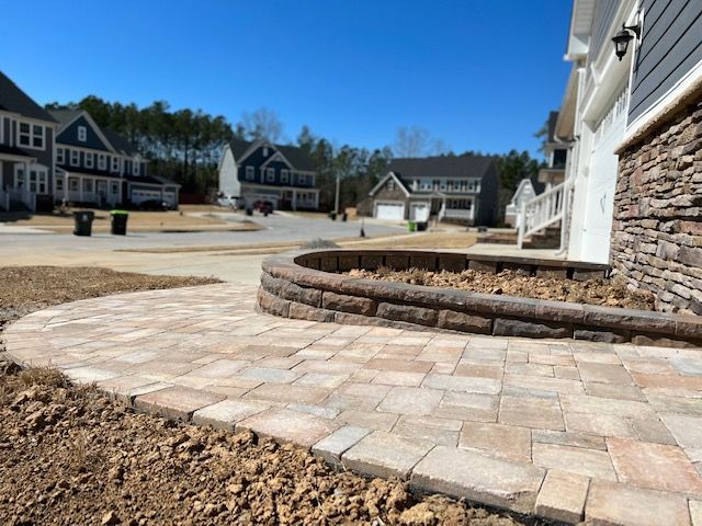 Paver walkway beside a stone house and curved retaining wall in a suburban neighborhood.