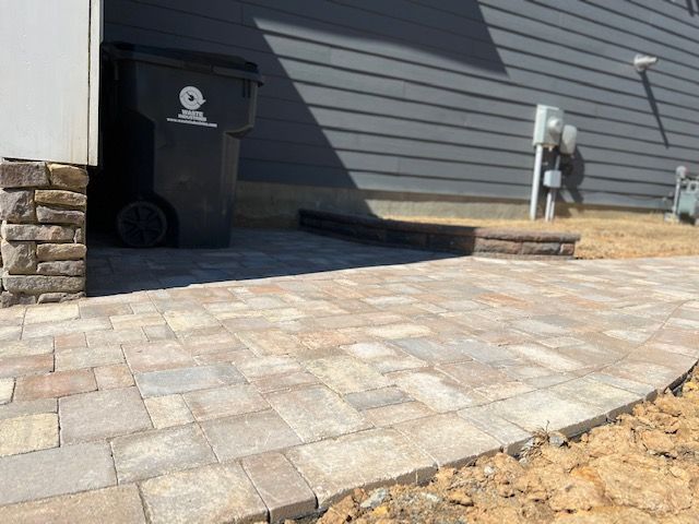 Brick-paved driveway beside a house with a black trash bin and utility meter