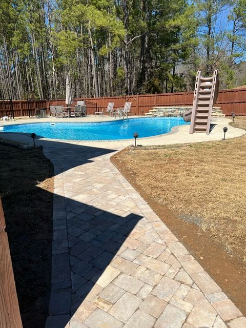 Backyard pool with stone patio, ladder, lounge chairs, and wooden fence under trees