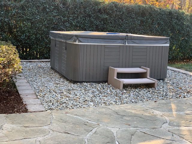 Outdoor hot tub on a gravel patio beside stone paving, with steps and a hedge backdrop